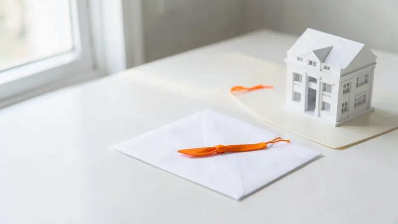Photograph of a white envelope with a loosening orange string beside a model office building