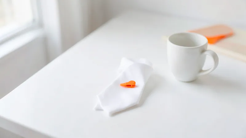 Photograph of a folded white necktie with an orange tie clip beside a white cup